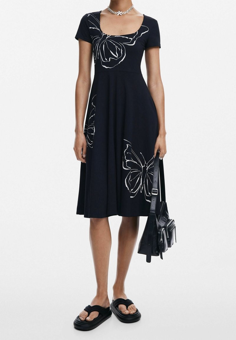 Une femme portant une robe noire mi-longue avec des imprimés papillons blancs, tenant un sac à main noir et portant des sandales à plateforme noires.