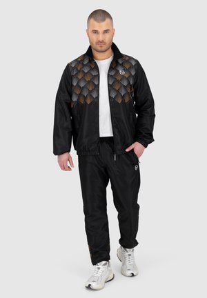 Giacca sportiva nera con un design a motivi in arancione, grigio e nero. Abbinata a pantaloni neri coordinati e scarpe da ginnastica bianche.