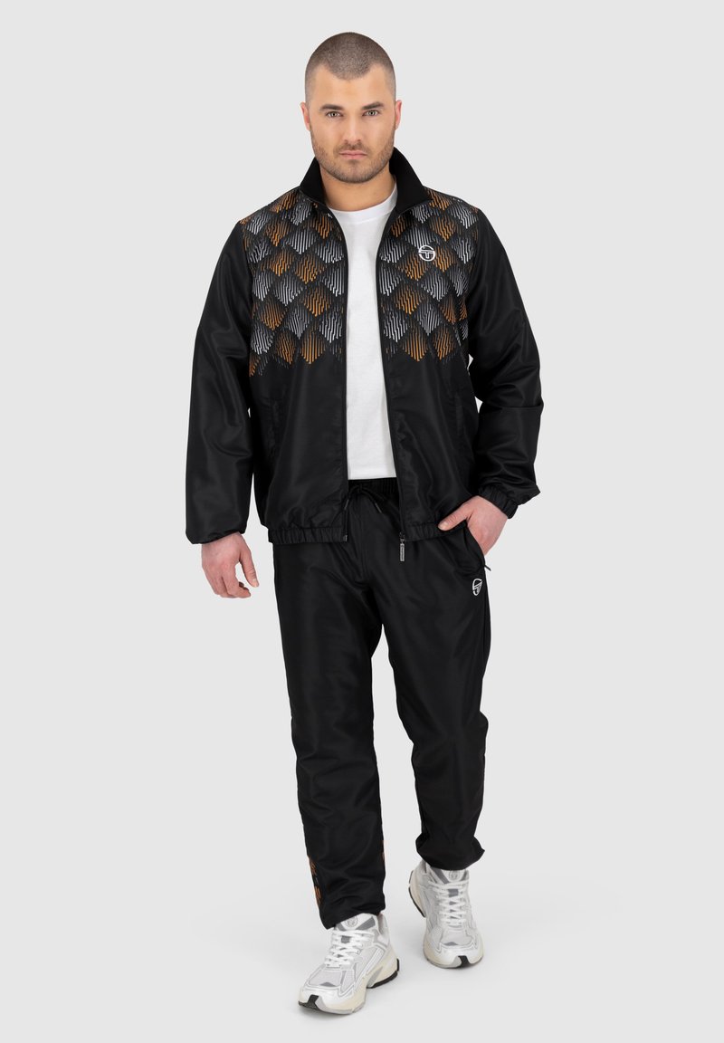 Zwarte sportjack met een patroonontwerp in oranje, grijs en zwart. Gepaarde met bijpassende zwarte broeken en witte sportschoenen.