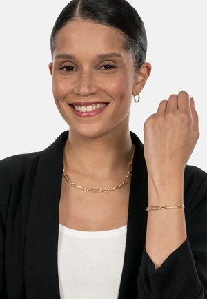 Schmuck in Goldtönen besteht aus einer Ketten- Halskette und einem Armband mit strukturierten Akzenten. Das Modell trägt ein schwarzes Blazer über einem weißen Oberteil.