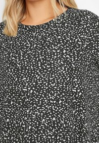 Svart tyg med vitt prickigt mönster, med en ribbad textur och rund halsringning. Designen är avslappnad och mångsidig.