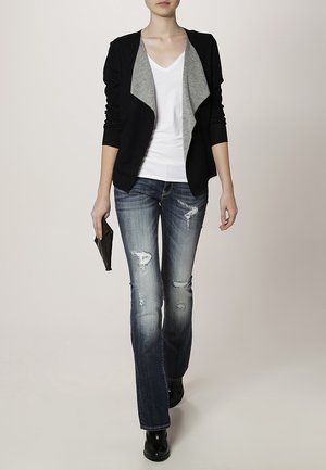 Femme portant une veste noire ouverte avec une doublure grise, un t-shirt blanc à col en V, un jean bleu déchiré, des bottes noires, et tenant une pochette noire.