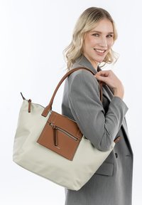 Beige tote bag with a brown leather front pocket, featuring a zip closure and two top handles. Material appears smooth and durable.