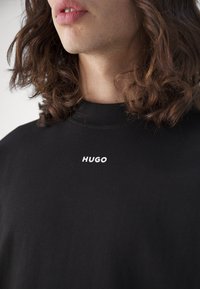Black t-shirt featuring a round neckline and short sleeves, with white "HUGO" text logo on the front, made from soft cotton fabric.