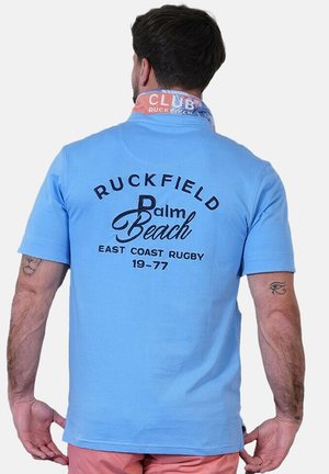 Homme portant un polo bleu clair avec l'inscription « Ruckfield Palm Beach East Coast Rugby 19-77 » au dos, un short rose et un foulard multicolore autour du cou.