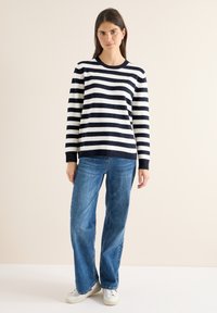 Laivastonsinivalkoraidallinen pehmeä neulepaita, jossa on pyöreä pääntie ja resorireunaiset hihat, yhdistetty vaaleansinisiin leveälahkeisiin farkkuihin ja valkoisiin lenkkareihin.