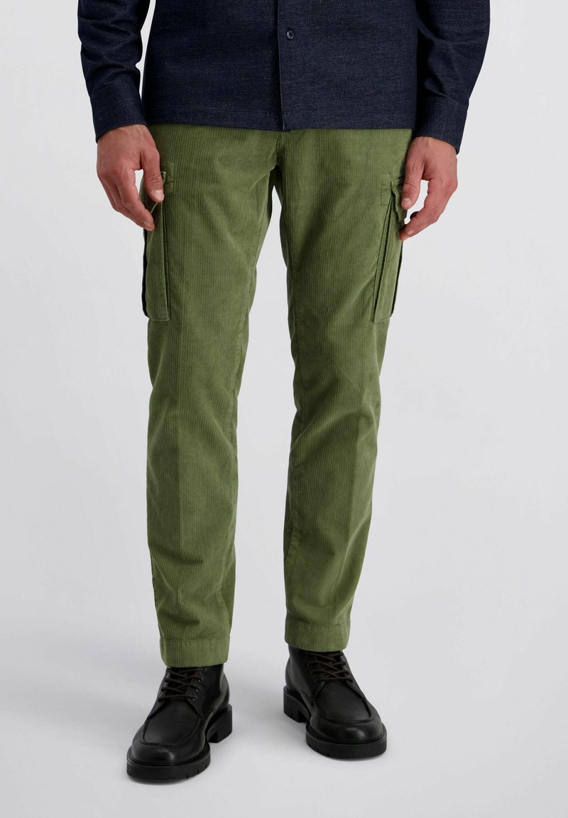 Man in olijfgroene corduroy cargobroek en zwarte leren veterschoenen, staande tegen een eenvoudige lichte achtergrond.