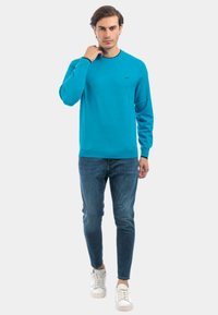 Felpa turchese a girocollo con maniche lunghe, abbinata a jeans skinny e scarpe da ginnastica bianche. Presenta un logo discreto sul petto.