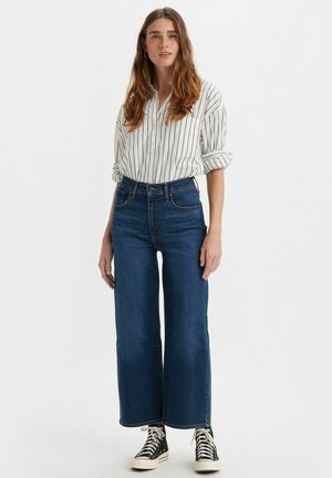 High-waisted wide-leg jeans in dark blue denim paired with a white striped long-sleeve shirt. Black high-top sneakers complete the look.