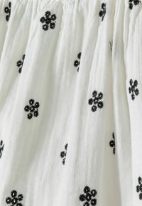 Tissu blanc avec une broderie florale noire, présentant un tissage léger et texturé ainsi que des détails froncés sur l'ensemble du matériau.