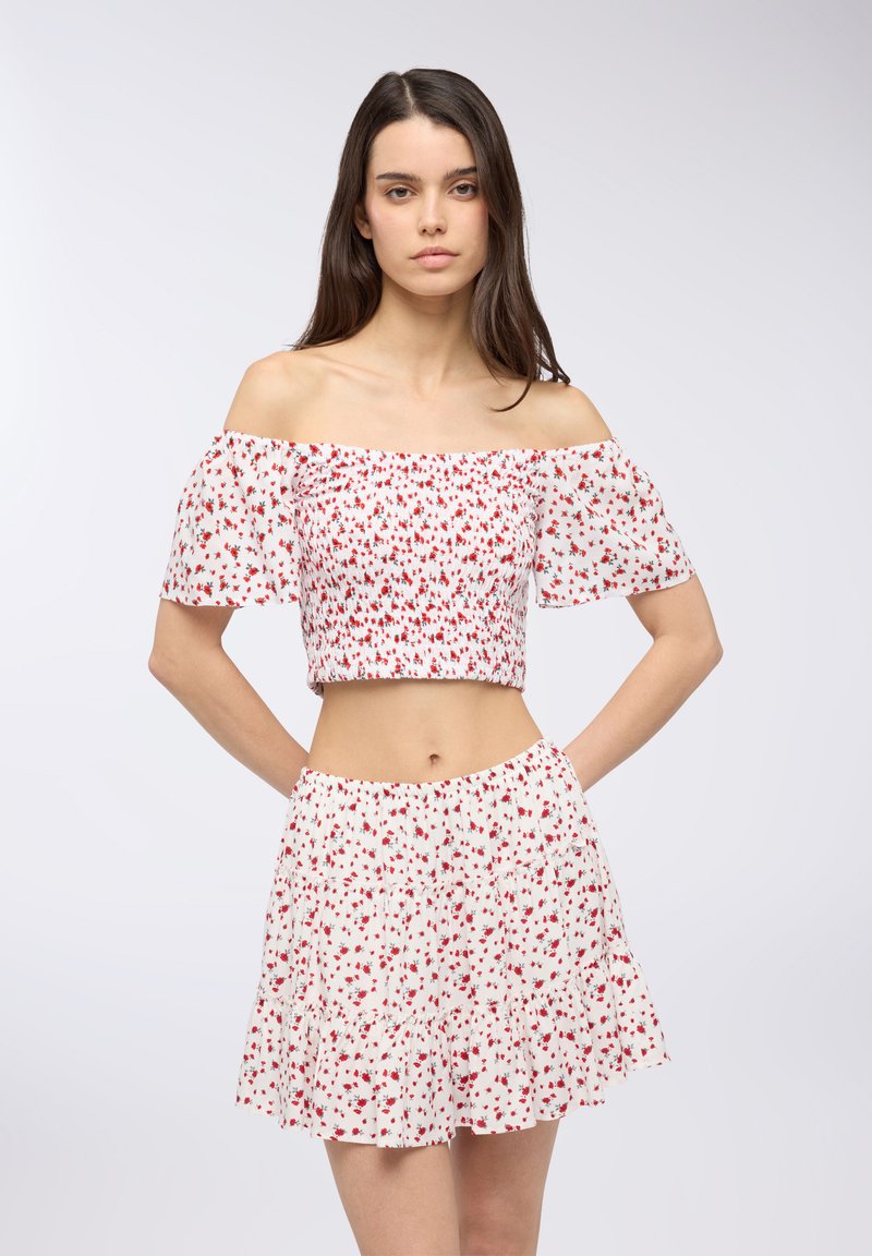 Crop top off-the-shoulder e gonna abbinata, entrambi bianchi con stampa floreale rossa. Presenta un design arricciato e una gonna a più strati con orlo arricciato.