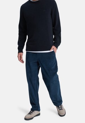 Man draagt een donkerblauwe trui, wit ondershirt, losse blauwe broek en beige sneakers met zwarte veters, staat met één hand in de zak.