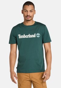 Muž v tmavě zeleném tričku s logem Timberland a světle hnědých cargo kalhotách, stojící na pozadí bílé zdi.