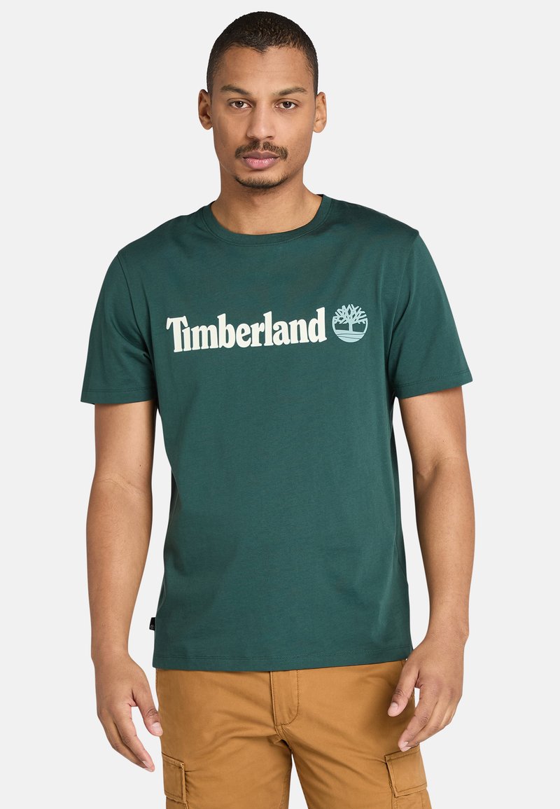 Muž v tmavě zeleném tričku s logem Timberland a světle hnědých cargo kalhotách, stojící na pozadí bílé zdi.