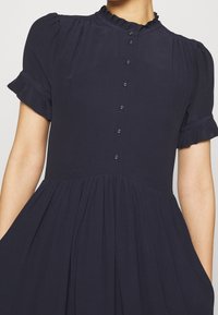 Granatowa sukienka zapinana na guziki z krótkimi bufkami, falbaniastym kołnierzykiem i zebranym w pasie. Wykonana z gładkiego materiału z lekko opadającą spódnicą.