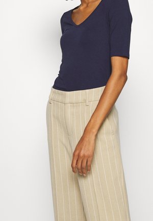 Vrouw die een marineblauw V-hals kortermige top draagt, ingestopt in beige pantalon met krijtstreep, staand met haar arm ontspannen langs haar zijde.