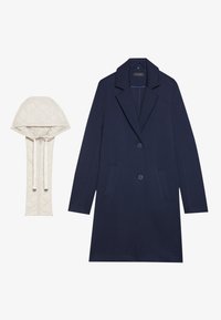 Cappotto lungo blu navy con due tasche e rever a punta, abbinato a un cappuccio rimovibile color crema imbottito con coulisse.