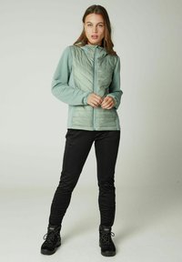 Mintgroene, gewatteerde jas met een capuchon, voorzien van verticale ribbels op de mouwen en een rits aan de voorkant, gecombineerd met zwarte broek en zwarte wandelschoenen.