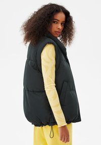 Black puffer vest with a high collar, side pockets, and cinched hem, worn over a yellow striped long-sleeve top. Textured, padded material.