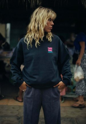 Młoda osoba o blond falowanych włosach stoi z rękami w kieszeniach, ubrana w ciemną bluzę z różowym i niebieskim napisem "SRF INC".