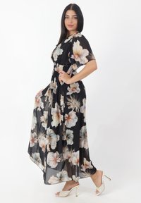 Robe maxi en tissu noir transparent ornée de grands motifs floraux dans des couleurs pastel, avec une taille smockée et des manches flottantes.