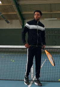 Svart velour treningsdrakt med hvite striper, jakke med glidelås og matchende bukser; holder en oransje tennisracket; innendørs banesetting.
