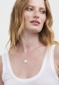 Srebrna ogrlica z obeskom v obliki kroga z detajlno zasnovo visi z nežne verižice, nošena čez bel top brez rokavov.
