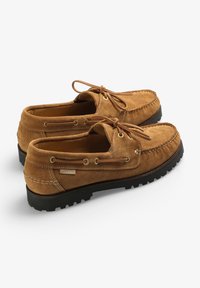 Des chaussures mocassins en daim beige avec des lacets marron, des œillets en métal, des surpiqûres blanches, des semelles en caoutchouc et une étiquette logo sur le côté.