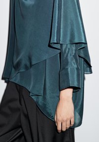 Satin top i teal med lagdelt design, har flydende ærmer og en rund kant, parret med sorte bukser. Glat tekstur synlig.