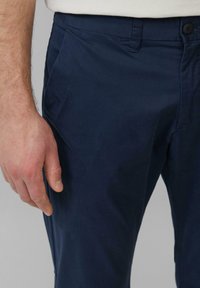 Männliche Hand, die neben einer marineblauen Hose mit einem geknöpften Bund und Gürtelschlaufen ruht, Hintergrund grau.