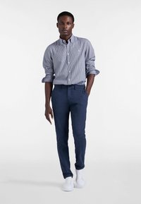 Camicia a maniche lunghe a bottoni con motivo a quadri blu e bianchi, abbinata a pantaloni navy e sneakers bianche. Tessuto morbido, vestibilità sartoriale e maniche arrotolate.