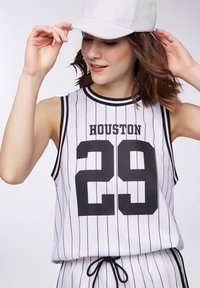 Canottiera bianca senza maniche con strisce verticali nere, con la scritta "HOUSTON" e il numero "29" in grassetto nero. Abbinata a un cappellino argento.