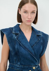 Gilet in denim di colore blu scuro, con un design strutturato delle spalle, rever affilati e dettagli di bottoni dorati in vita, caratterizzato da cuciture visibili.