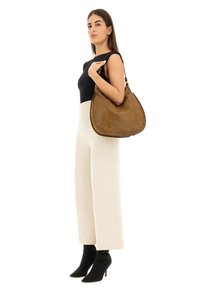 Borsa hobo in pelle marrone con design floreale inciso, dotata di forma rotonda, texture morbida e tracolla. Modello che indossa una maglia nera e pantaloni bianchi.