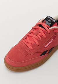 Sapatilha de camurça cor-de-rosa coral com detalhes em preto, biqueira perfurada e sola de borracha castanha com aderência texturizada. Marca Reebok na língua.