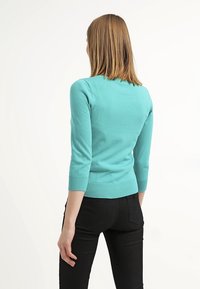 Tyrkosa tröja med trekvartsärmar, ribbat fåll och slät textur. Bärs med svarta byxor; bakvy är synlig.