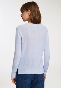 Kobieta z brązowymi włosami do ramion, ubraną w jasnoniebieski sweter dzianinowy i ciemnoniebieskie dżinsy, stoi plecami do prostego tła.