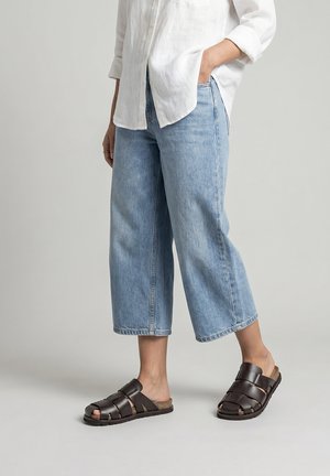 Personne portant un jean bleu clair court, une chemise blanche à manches longues et des sandales ouvertes en cuir marron foncé, debout avec une main dans la poche.