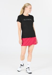 Kvinna iförd svart FZ Forza t-shirt, röd sportkjol, vita strumpor och vita sneakers, stående och tittande åt sidan med ett leende.