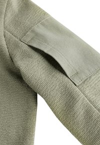Olivzaļas krāsas džempera piedurkne ar teksturētu adījuma audumu, gludiem akcentiem un ielāpu kabatu augšējā rokā.
