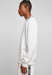 Hvid sweatshirt med rund halsudskæring, prydet med sort brandbeslag. Lange ærmer og løs pasform med ribstrikkede manchetter og kant.