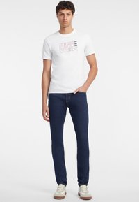 Balta medvilninė marškinėliai su "GUESS JEANS" grafika, derinami su tamsiai mėlynomis džinsais ir baltais sportbačiais su kreminiais akcentais.