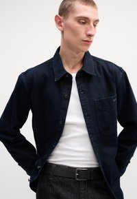 Camisa de ganga escura com um design de botões, detalhe de bolso e costura em contraste, usada sobre uma t-shirt branca de alças e combinada com um cinto preto.