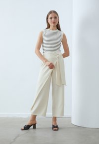 Sleeveless striped top in black and white paired with cream wide-leg trousers featuring a front wrap and tie detail. Black heeled sandals.