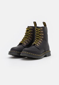Botas de cuero negras hasta el tobillo con cordones amarillos, con ojales de metal y una suela cosida en amarillo. Incluye una tira de agarre en la lengüeta.