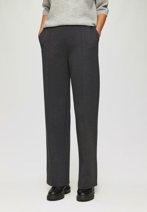 Persoon gekleed in donkergrijze rechte broek, zwarte chunky schoenen en een lichtgrijze trui met de handen in de zakken, staand tegen een neutrale achtergrond.