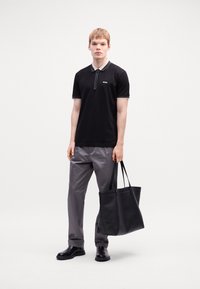 Juodas polo marškinėliai su pilku apykakle, derinami su pilkomis kelnėmis. Modelis laiko juodą pirkinių maišą. Lygus audinys, modernus fasonas, minimalus prekės ženklas.