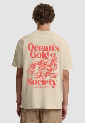 Beżowy t-shirt z czerwonym nadrukiem homara, plasterkiem cytryny i napisem „Ocean’s Gold Society”, noszony przez osobę o kręconych brązowych włosach, stojącą tyłem do kamery.