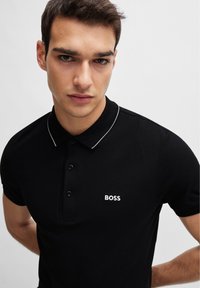 Svart polo-skjorte med krage, med en tre-knappers knappestolpe og hvit brodert "BOSS"-logo på brystet. Glatt tekstur.