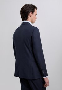 Blazer azul marino, corte entallado, abertura simple en la parte posterior, tejido ligero, camisa de vestir azul claro a contraste visible en el cuello.